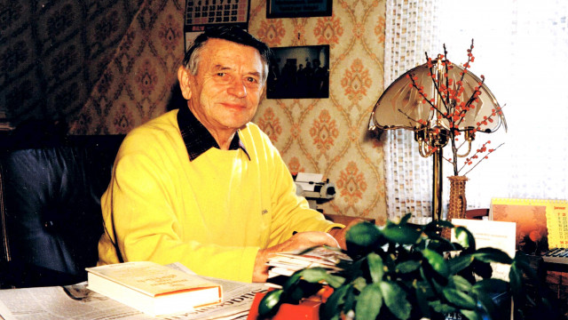 Heiner Schneier sitzt in einem gelben Pullover hinter seinem Schreibtisch und blickt in die Kamera.